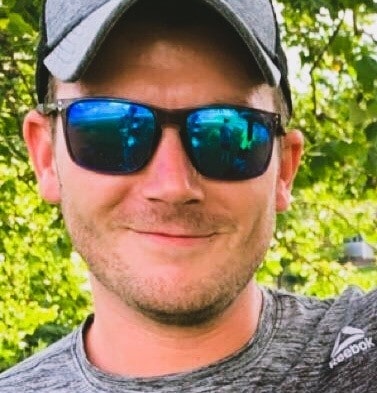 Male wearing sunglasses and hat in front of trees